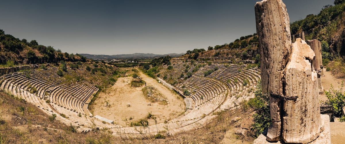 An ancient amphitheater with tiered seating and a central stage, surrounded by hills and vegetation.