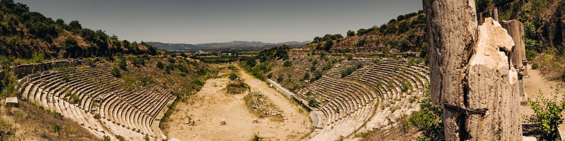 An ancient amphitheater with tiered seating and a central stage, surrounded by hills and vegetation.