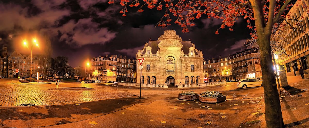 A historic building with intricate architecture, a cobblestone plaza, and a tree with autumn leaves.