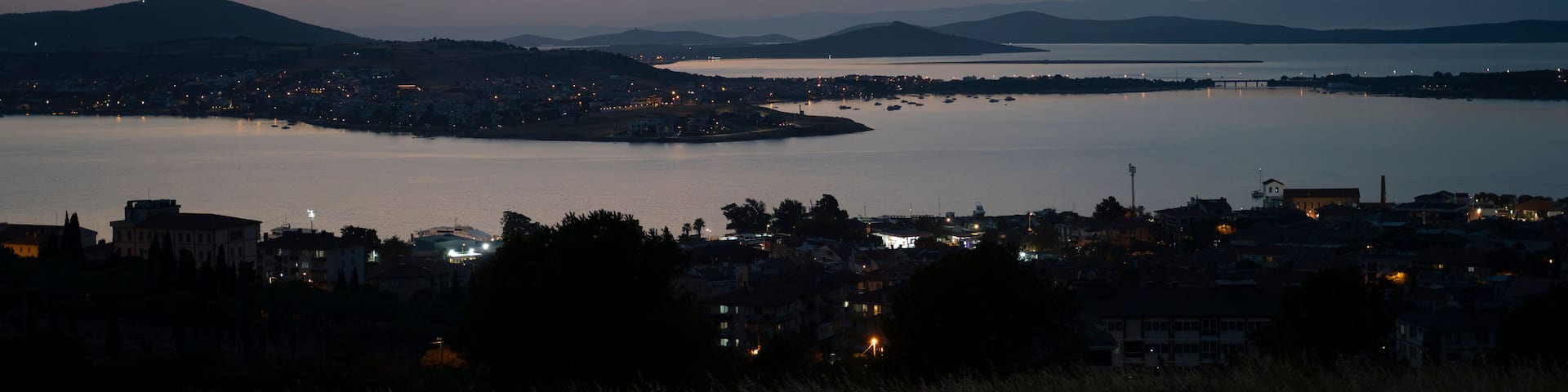 A coastal cityscape at dusk with a large body of water, a bridge, and distant mountains.