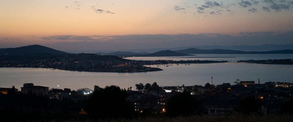 A coastal cityscape at dusk with a large body of water, a bridge, and distant mountains.