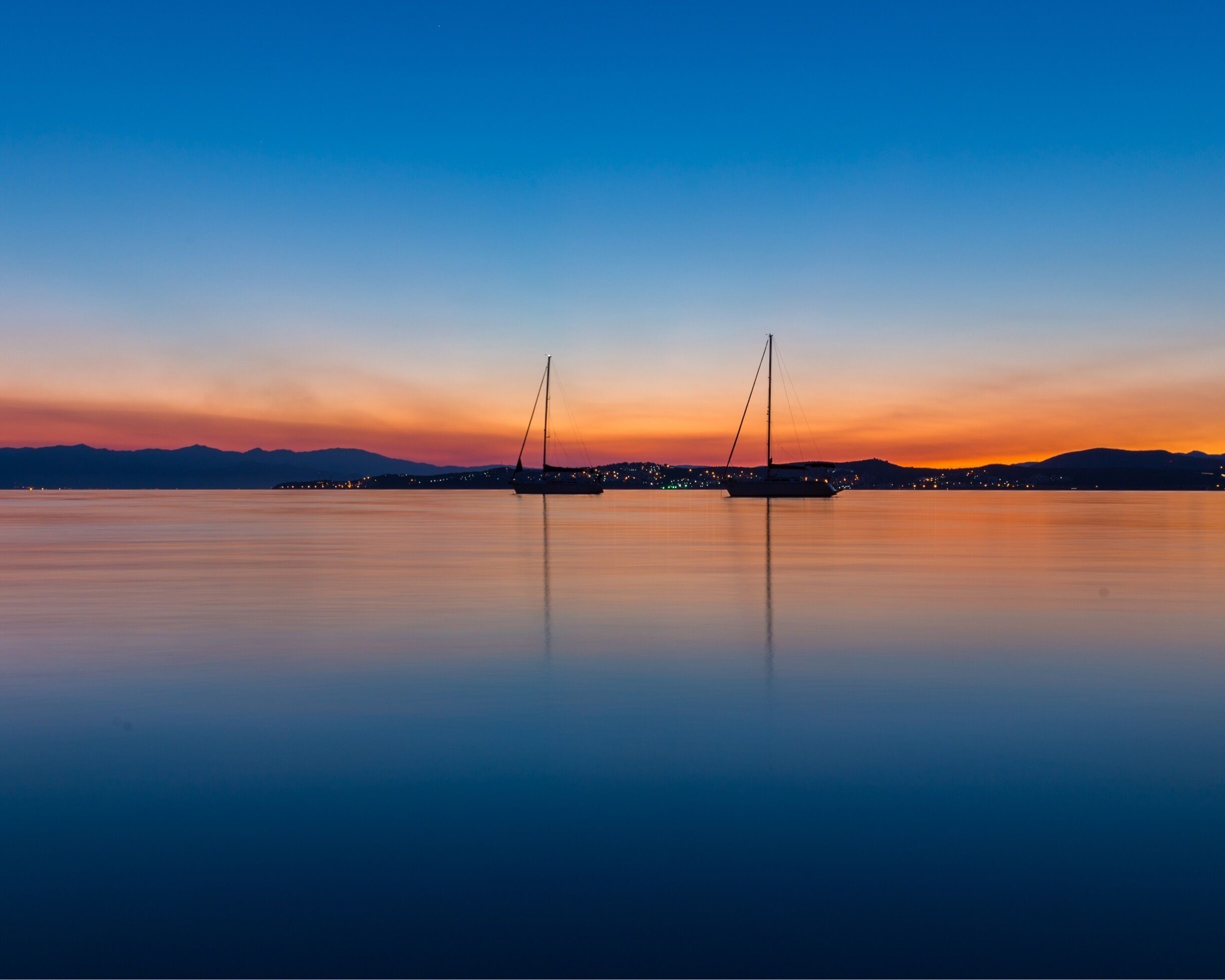 A calm lake at sunset with two sailboats anchored in the center, surrounded by a city skyline with lights reflecting on the water.
