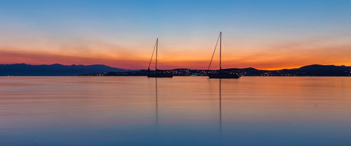 A calm lake at sunset with two sailboats anchored in the center, surrounded by a city skyline with lights reflecting on the water.