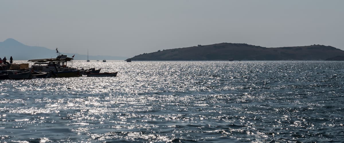A calm sea with boats docked on the shore and a distant hill.