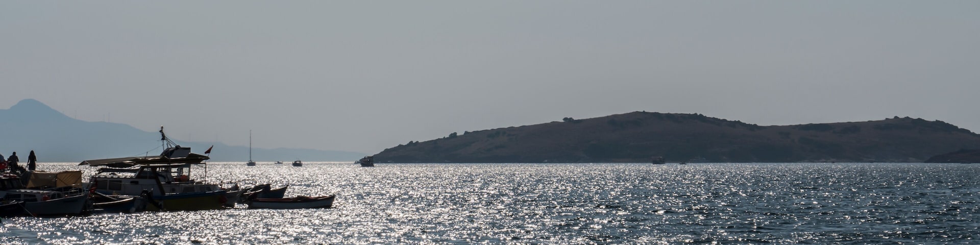 A calm sea with boats docked on the shore and a distant hill.