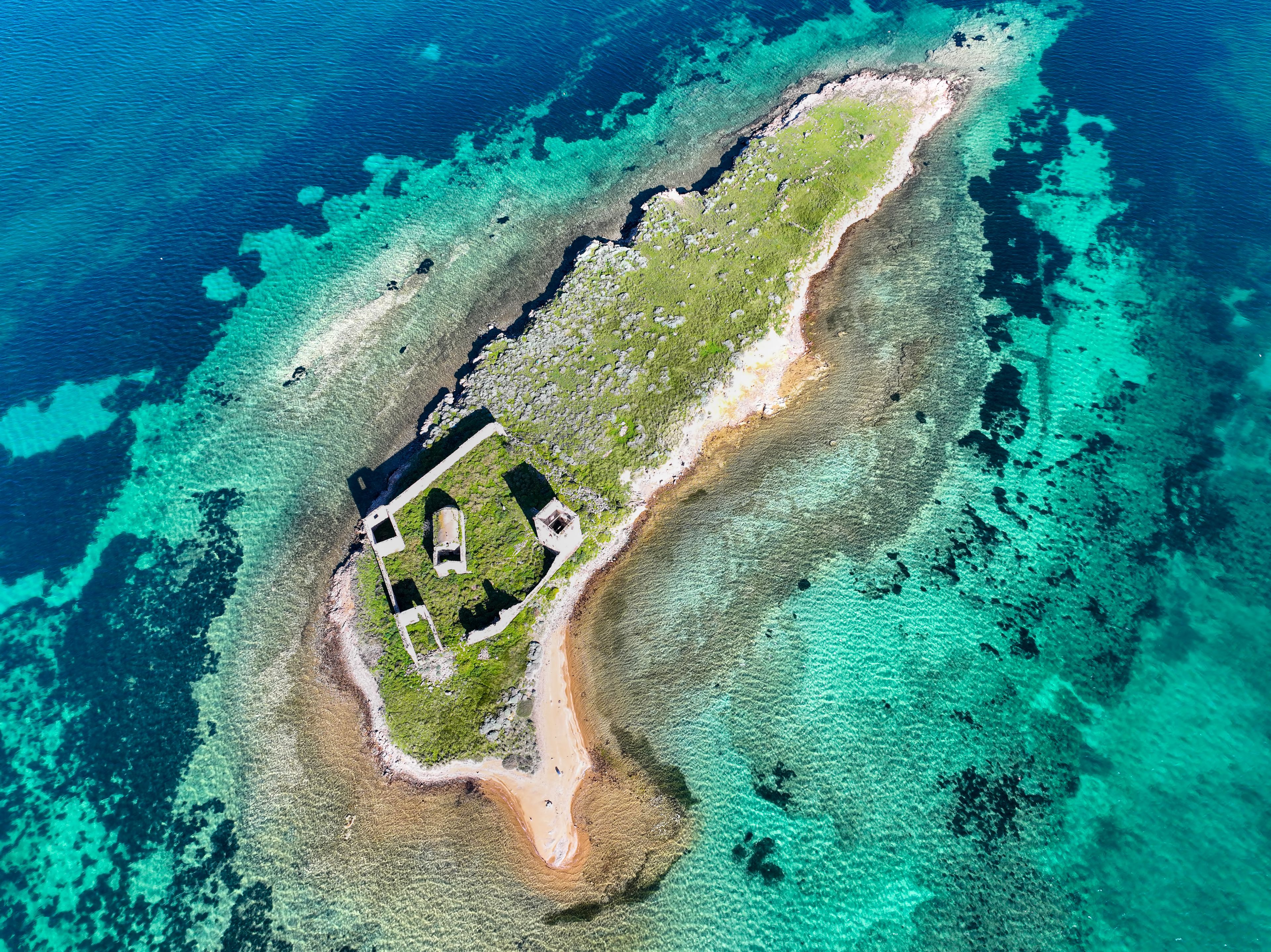 Vista aérea de una pequeña isla con aguas turquesas cristalinas, una playa arenosa y algunas construcciones.