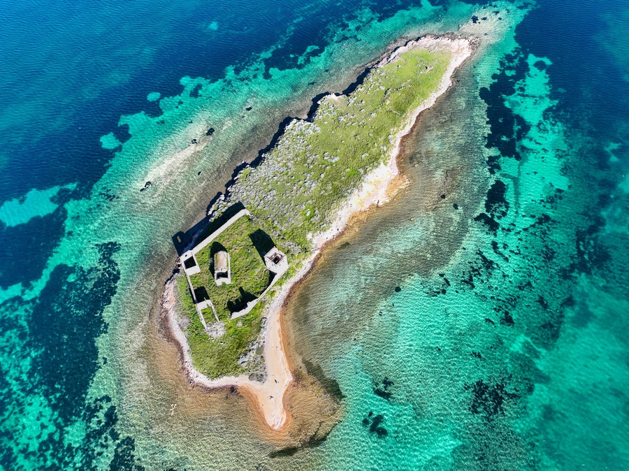 Vista aérea de una pequeña isla con aguas turquesas cristalinas, una playa arenosa y algunas construcciones.