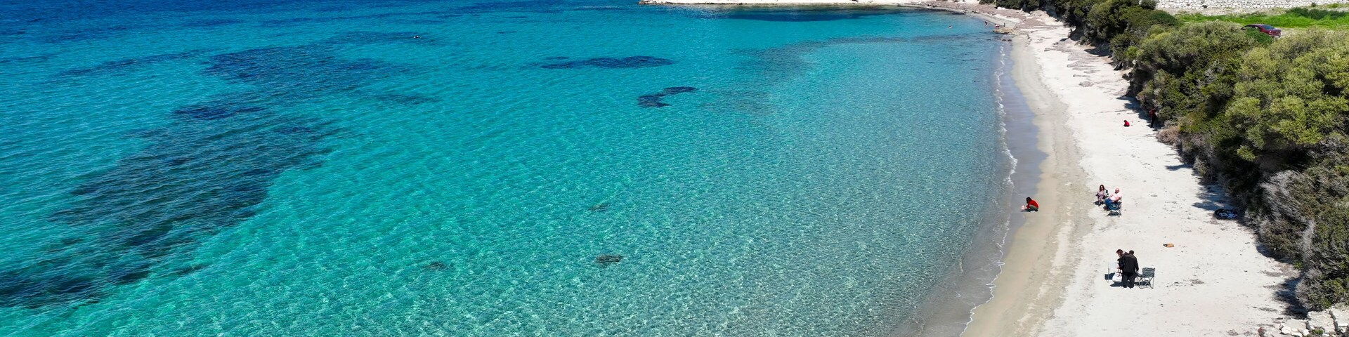 Un mar turquesa transparente, una playa de arena y un paisaje montañoso con vegetación escasa.