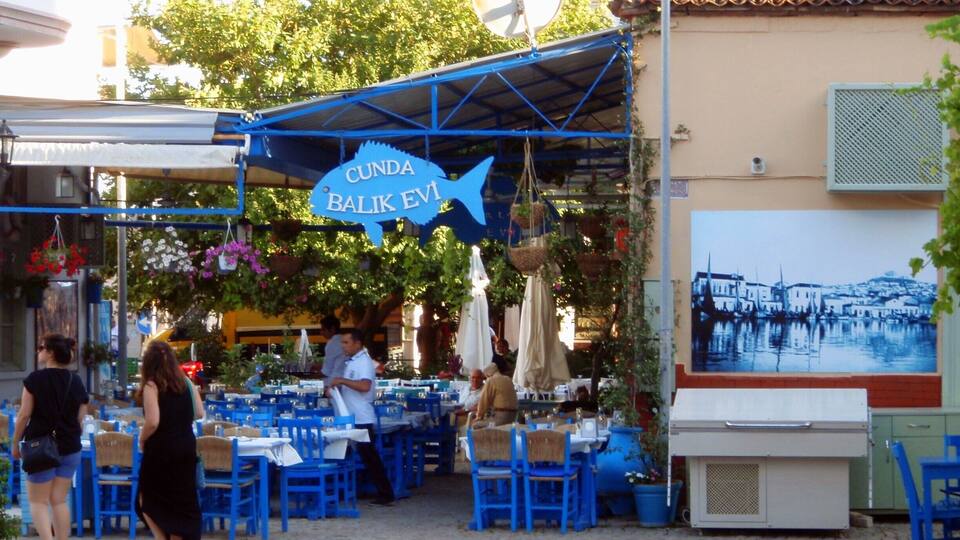 A seaside restaurant with blue tables and chairs, a sign reading "Cunda Balik Evi", and a mural of a coastal scene.