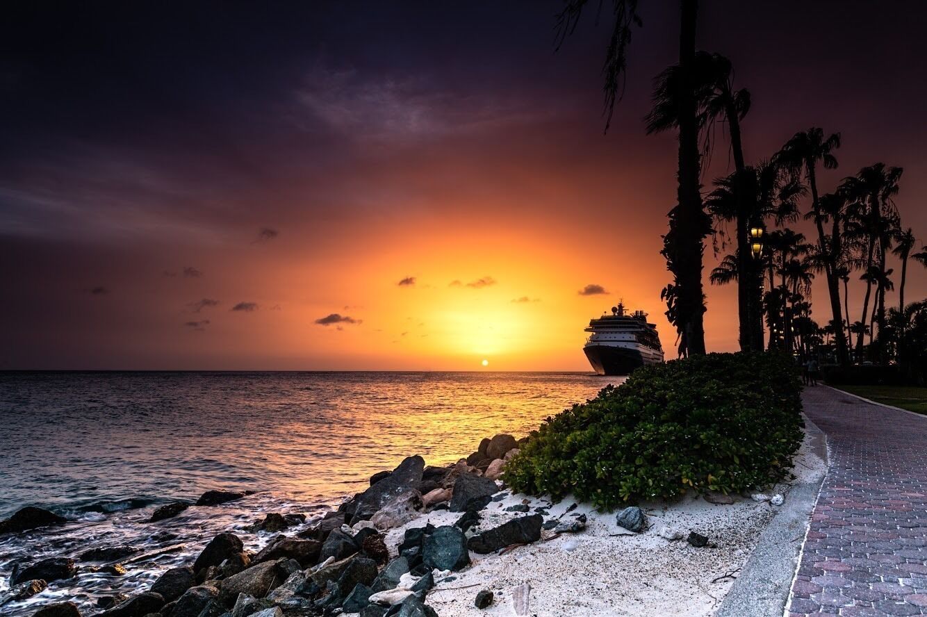A sunset over the ocean with a cruise ship, palm trees, and a paved walkway.