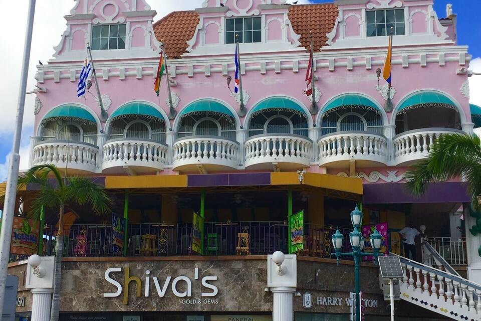 Ein pink-weißes Gebäude mit mehreren Flaggen, einem Schild für 'Siva's Gold & Gems' und einer Treppe, die zum Eingang führt.