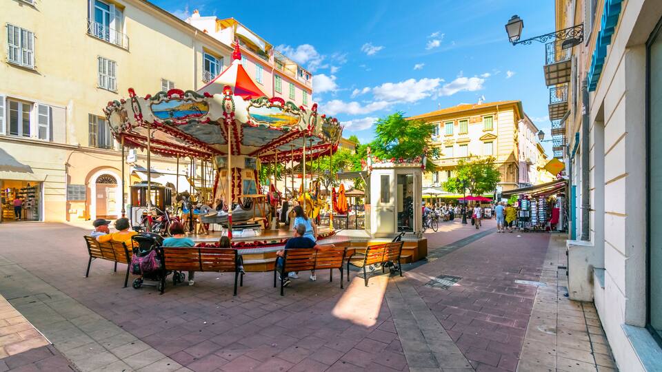 A carousel in a square with benches, surrounded by buildings with balconies and shops.