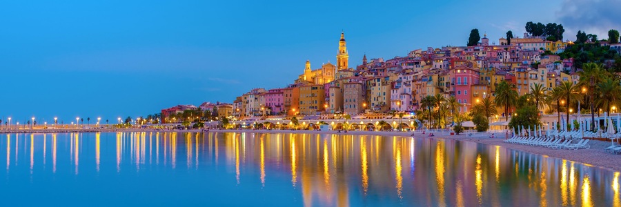 Un paisaje urbano costero al atardecer con una torre destacada, edificios coloridos y un mar tranquilo que refleja luces.