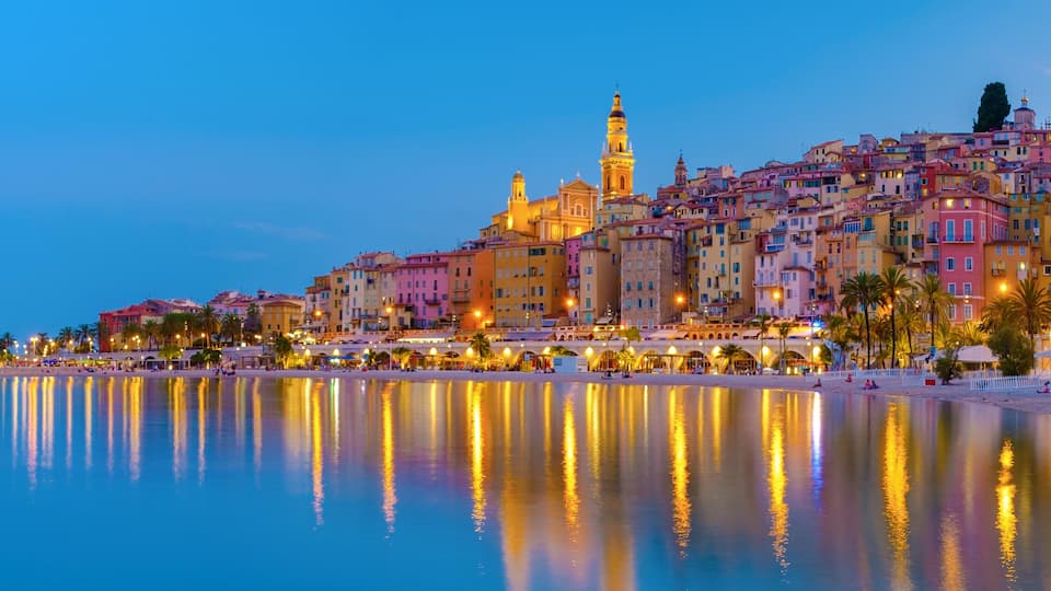 A coastal cityscape at dusk with a prominent tower, colorful buildings, and a calm sea reflecting lights.