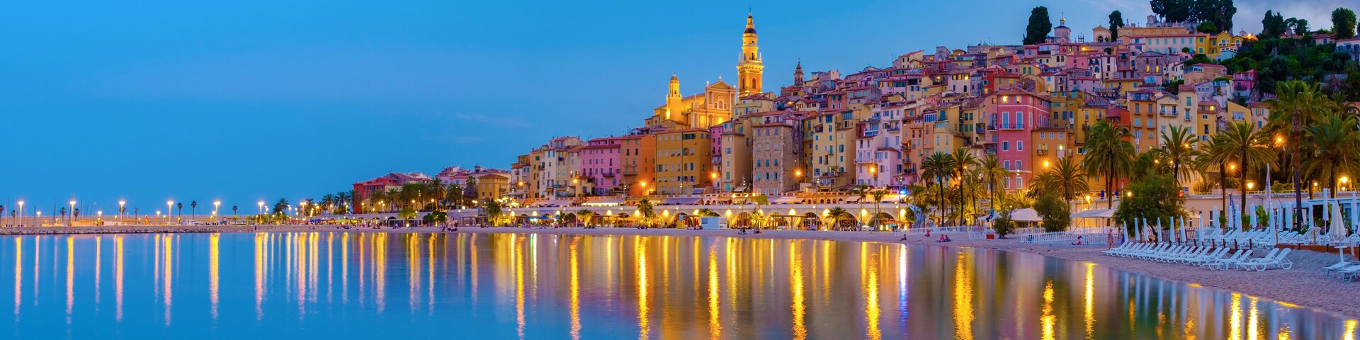 A coastal cityscape at dusk with a prominent tower, colorful buildings, and a calm sea reflecting lights.
