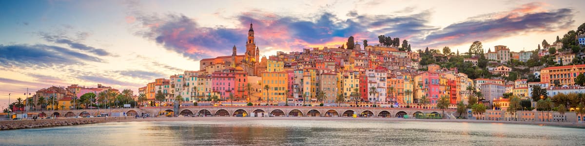 A coastal cityscape with colorful buildings, a prominent tower, and a bridge over the water.