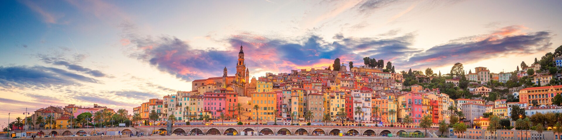 Un paisaje urbano costero con edificios coloridos, una torre destacada y un puente sobre el agua.