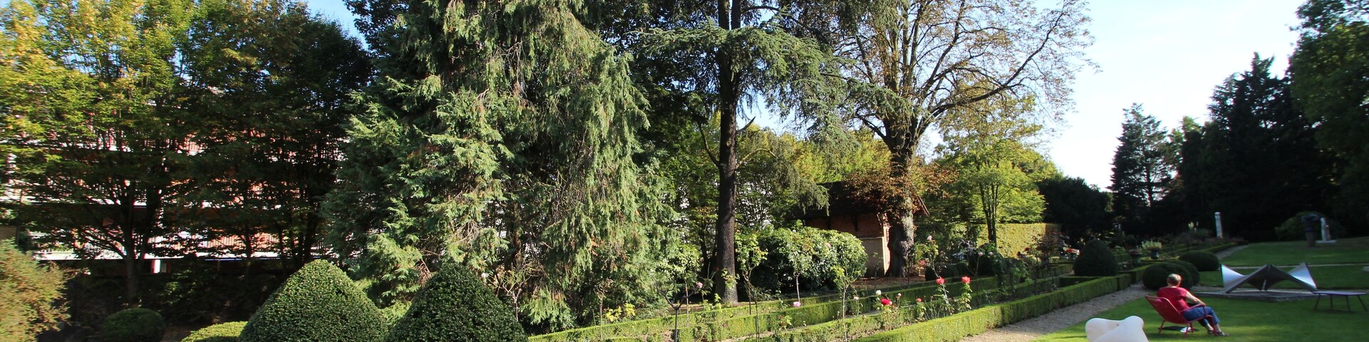 Um jardim bem cuidado com sebes aparadas, uma árvore grande e uma escultura em um pedestal.