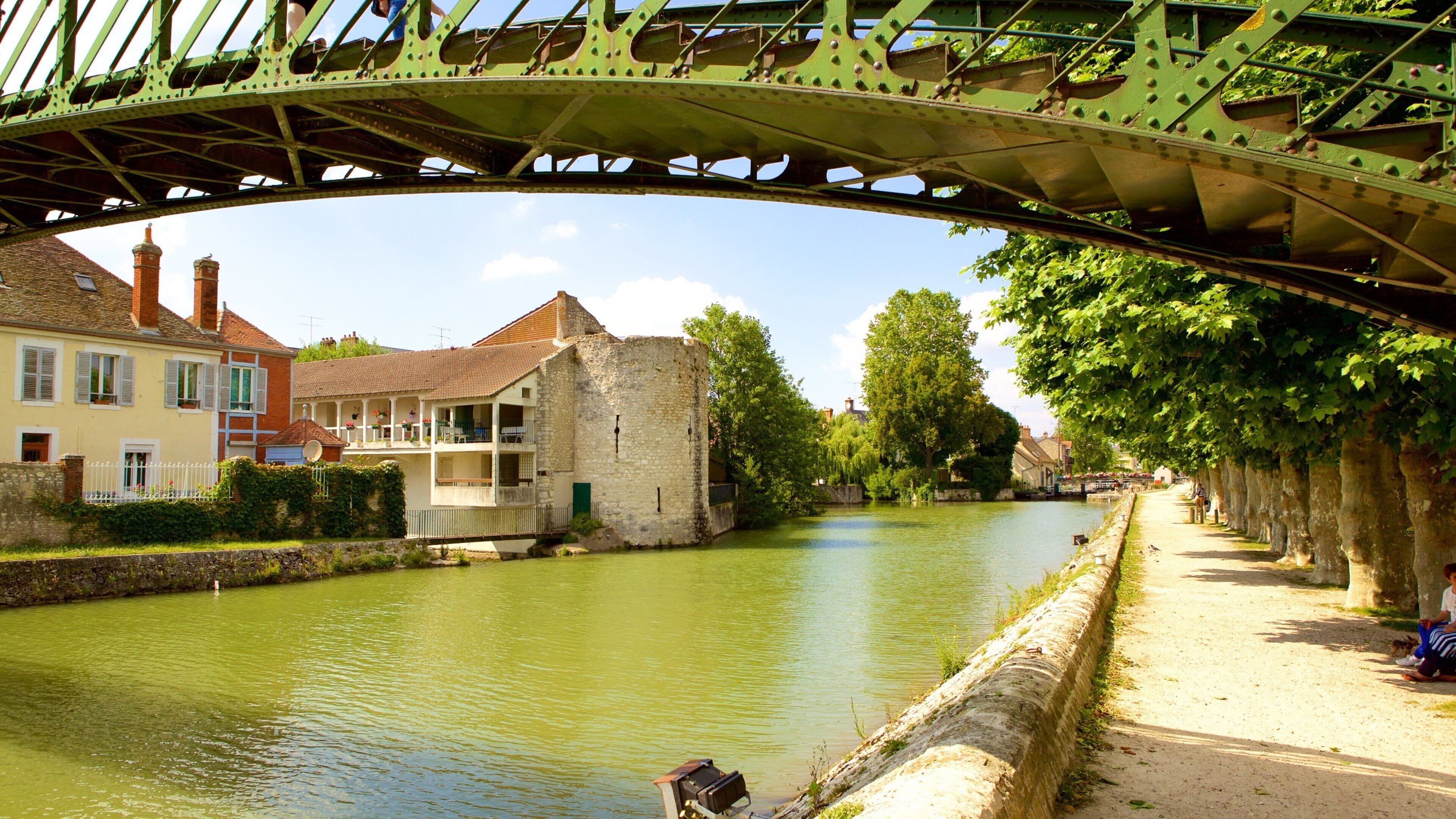 A green bridge over a river, stone buildings, and a tree-lined path.