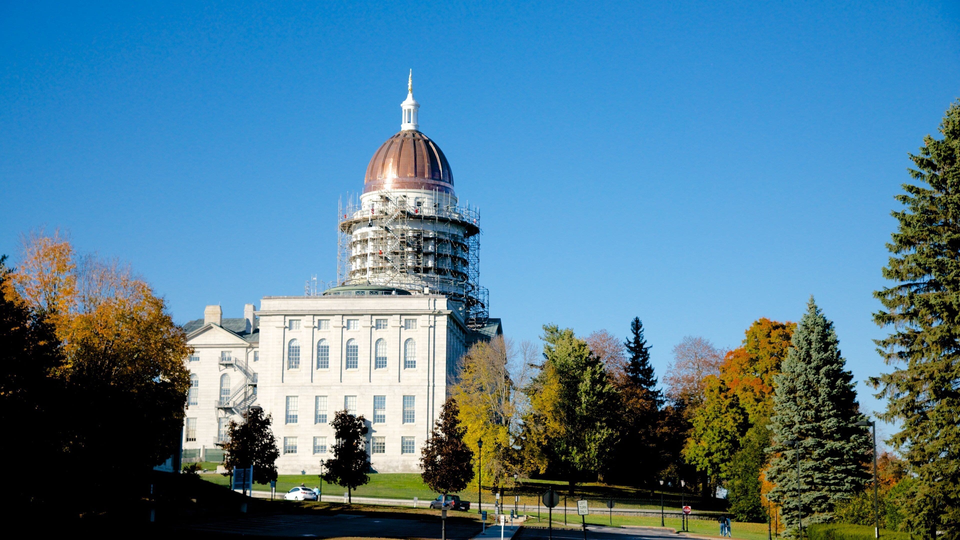 A historic building with a dome and scaffolding, surrounded by autumn trees and a clear blue sky.