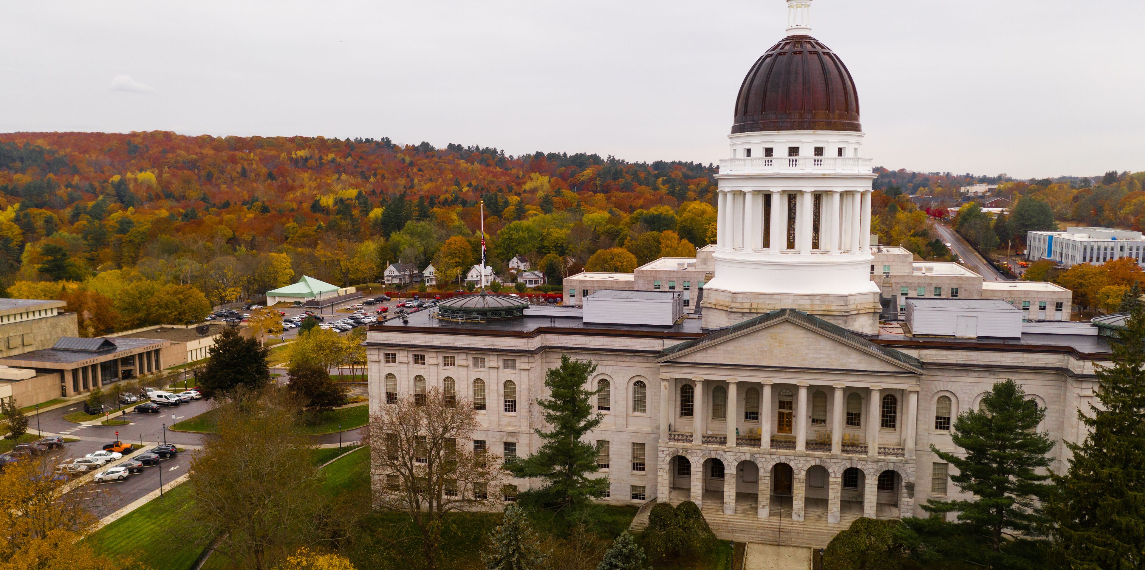 A large white building with a dome and columns, surrounded by autumn-colored trees and a hilly landscape.