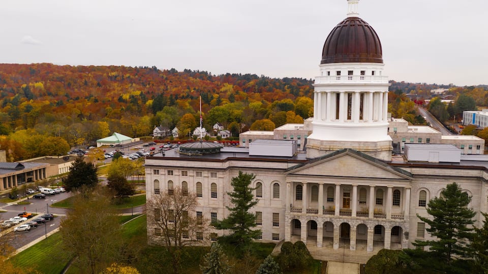 A large white building with a dome and columns, surrounded by autumn-colored trees and a hilly landscape.