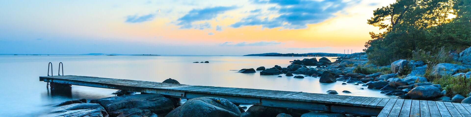 A wooden pier extending into calm waters with large rocks and a sunset in the background.