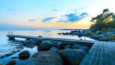 Un muelle de madera que se adentra en aguas tranquilas, con grandes rocas y un atardecer de fondo.