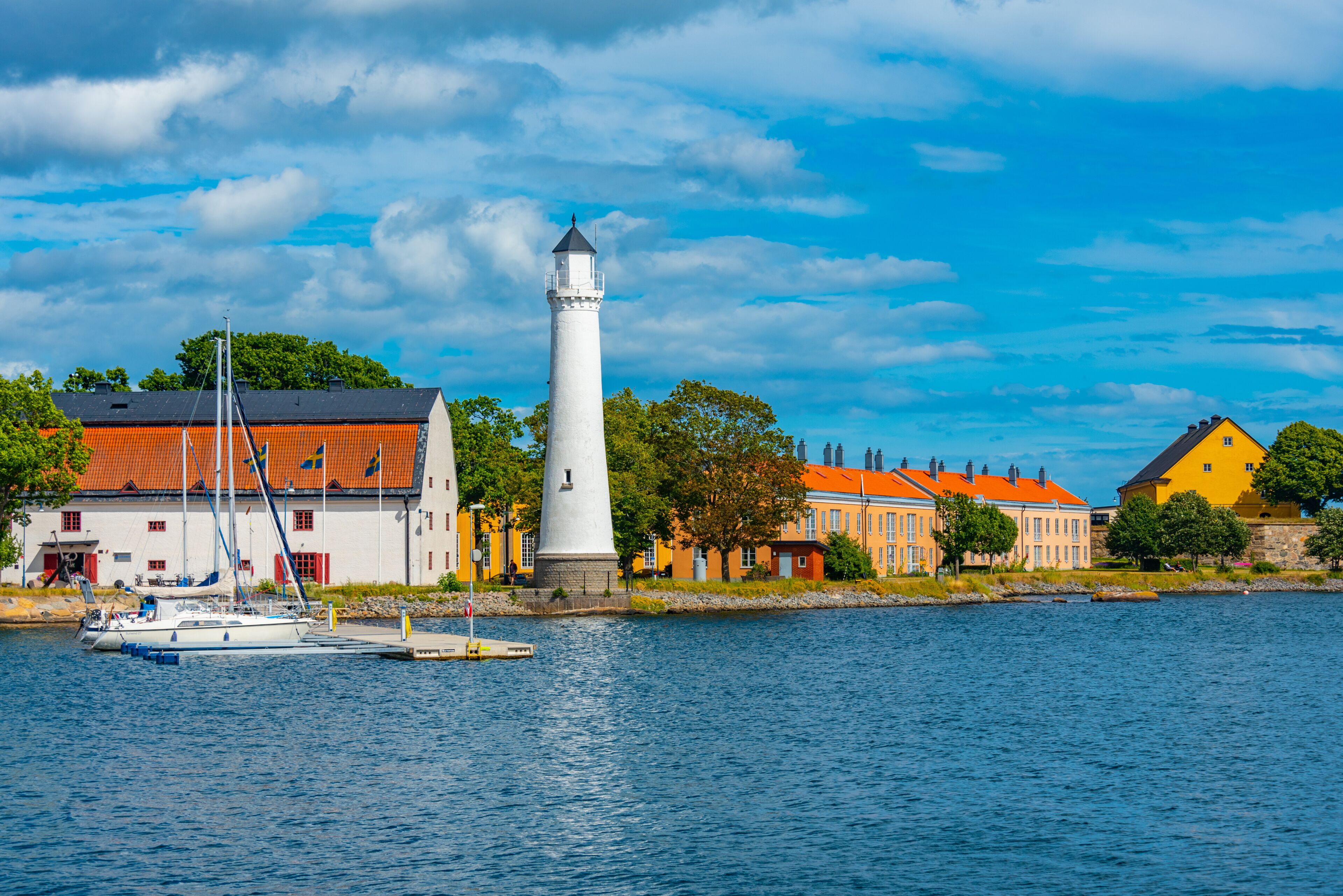A lighthouse, a white building with a red roof, and a yellow building with a red roof by a body of water.