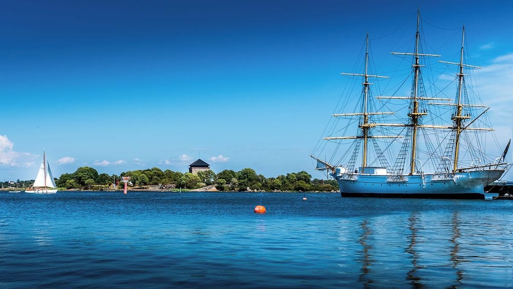 Ein großes Segelschiff liegt an der Küste, im Hintergrund sind ein kleines Segelboot und ein Gebäude zu sehen.