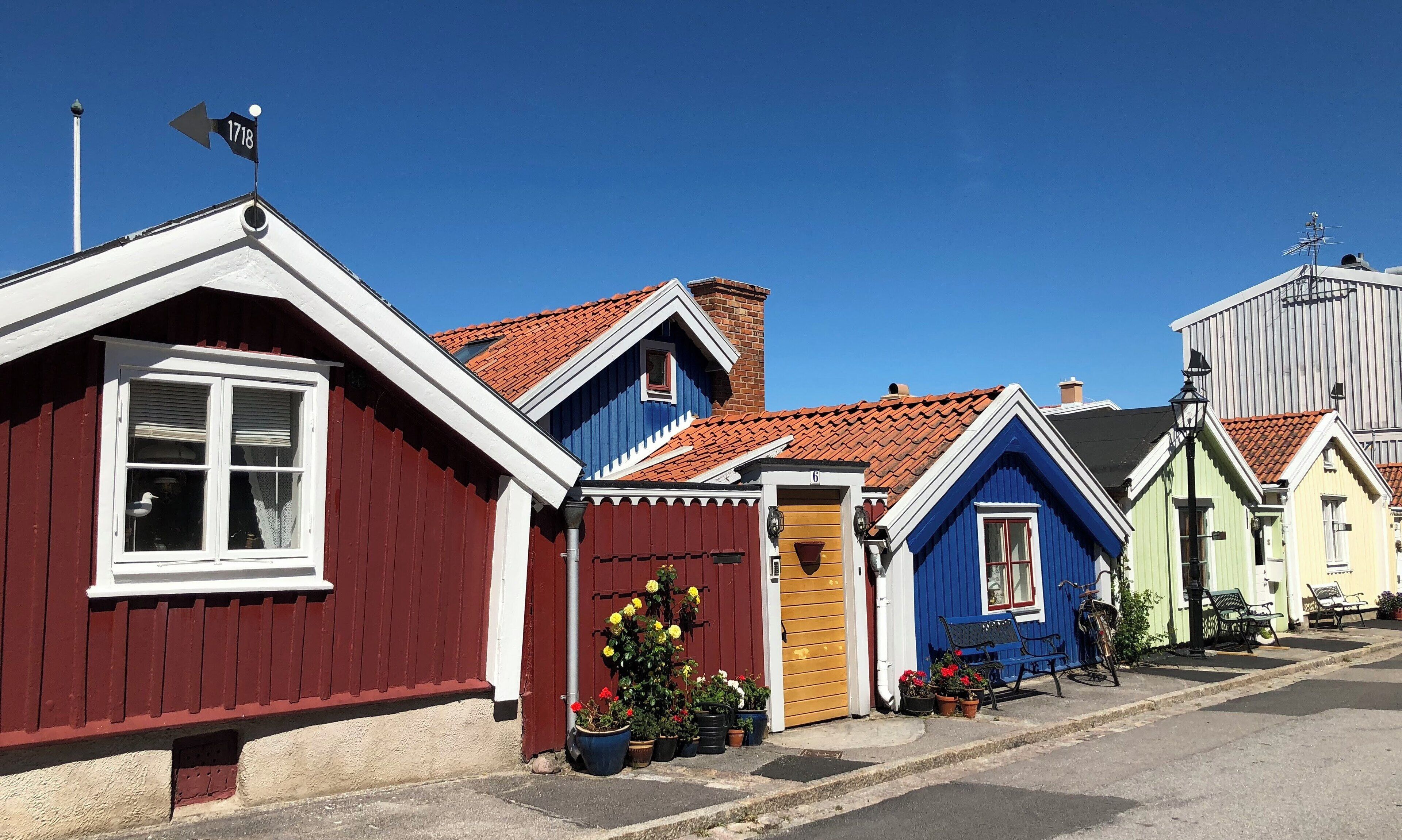 A row of colorful houses with red, blue, and yellow facades, a clear blue sky, and a street with benches and potted plants.