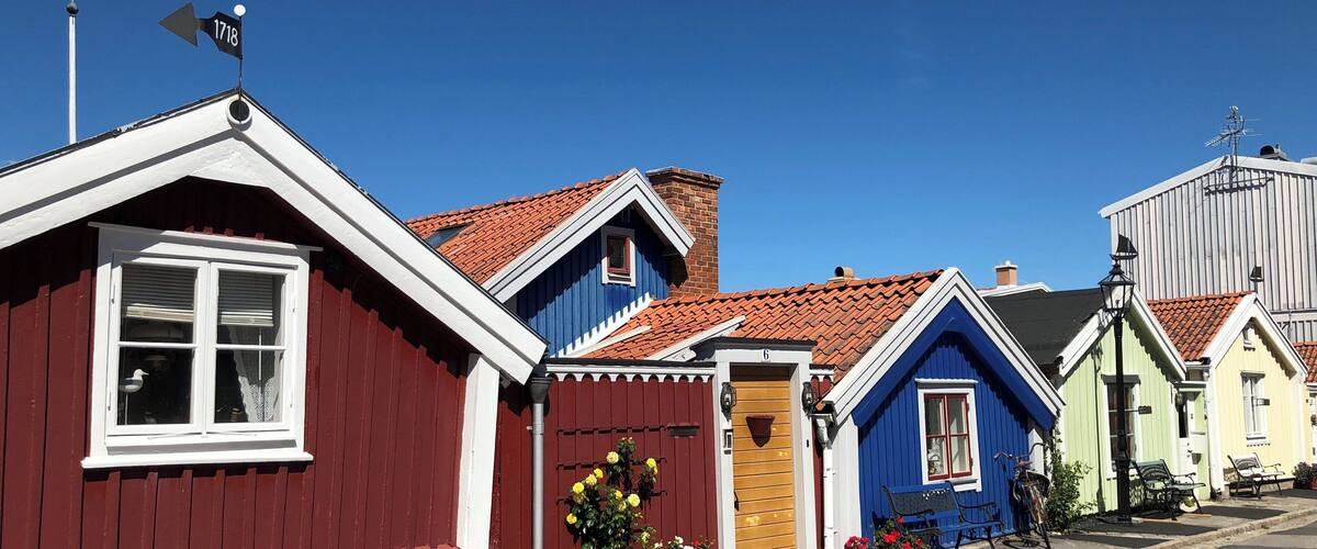 A row of colorful houses with red, blue, and yellow facades, a clear blue sky, and a street with benches and potted plants.