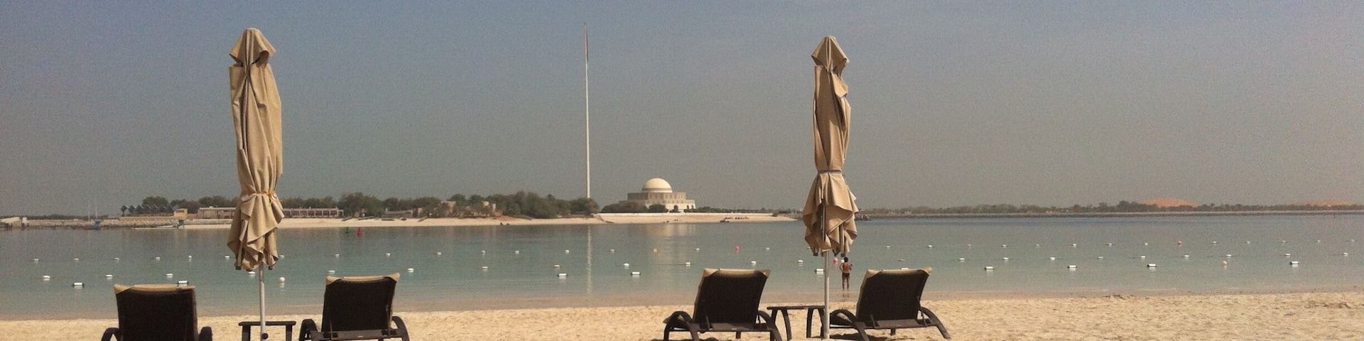 Une plage avec trois chaises longues et deux parasols, un ciel dégagé et, au loin, une structure en forme de dôme blanc.