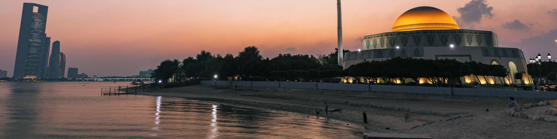 A sunset over a body of water with a modern building and a dome structure with a flag.