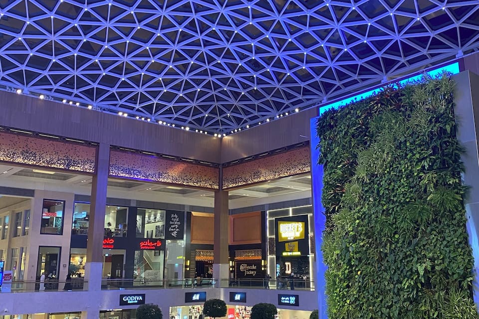 A modern shopping mall with a geometric ceiling design, a green vertical garden, and various retail stores.
