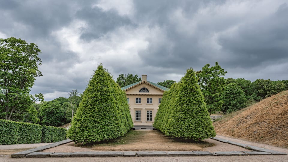 A stately building with a symmetrical facade, flanked by two tall, conical trees, under a cloudy sky.
