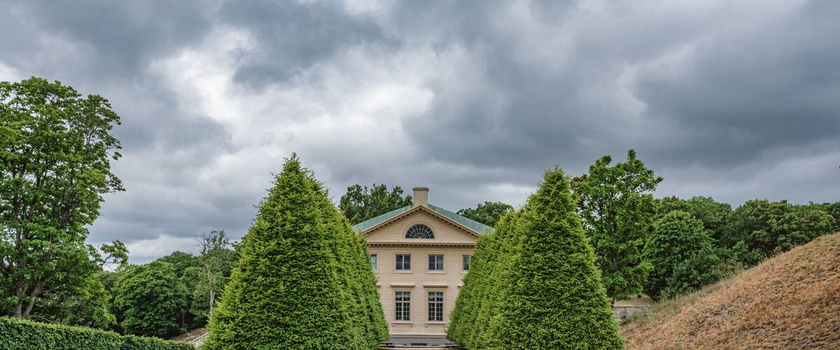 A stately building with a symmetrical facade, flanked by two tall, conical trees, under a cloudy sky.
