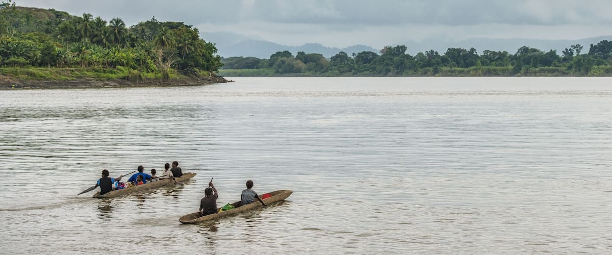 A group of people in a longboat on a calm river, surrounded by lush greenery and distant hills.