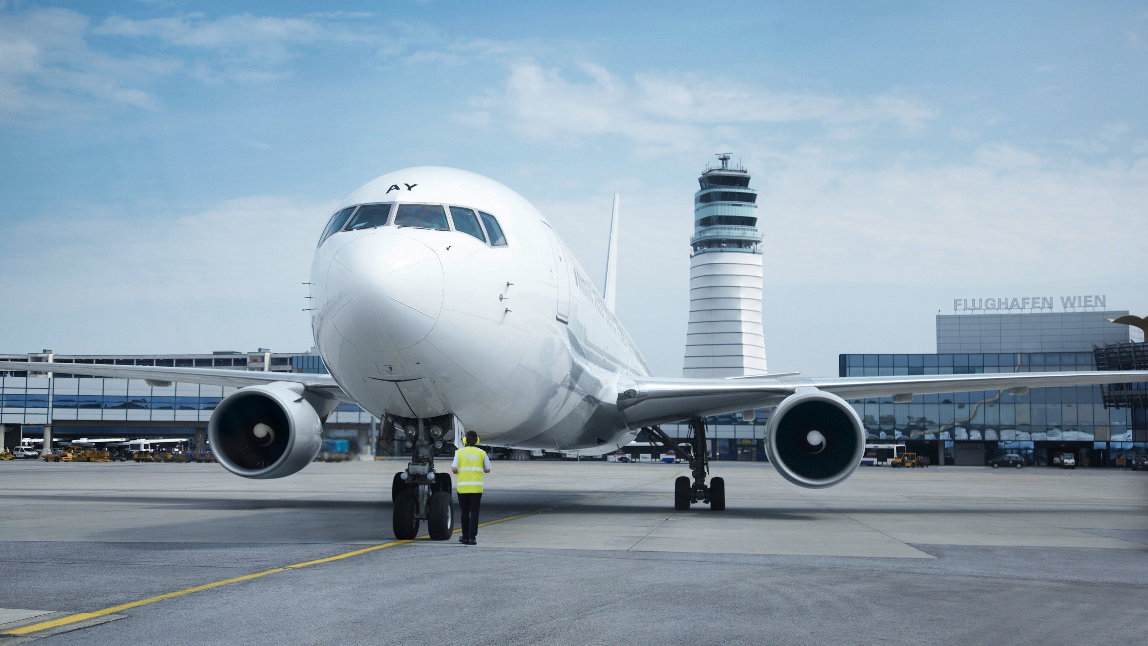 An airport with a large white airplane parked on the tarmac, a control tower, and the building labeled "FLUGHAFFEN WIEN" in the background.