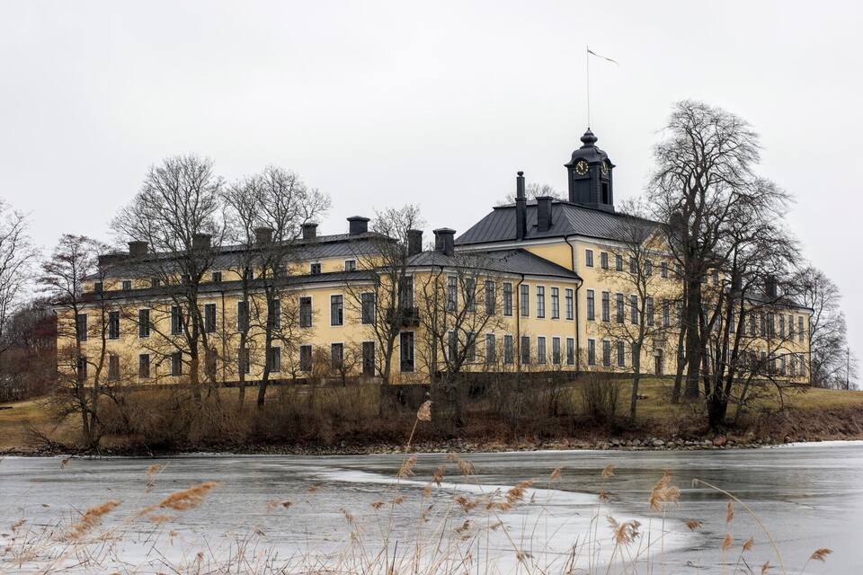 A large yellow building with a clock tower, surrounded by bare trees and a frozen river.
