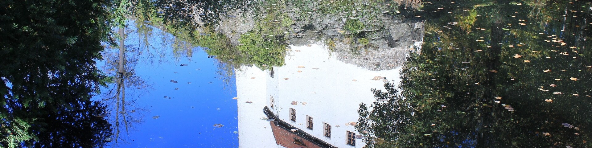 A calm river reflecting a white building with a tower, surrounded by dense foliage and trees.