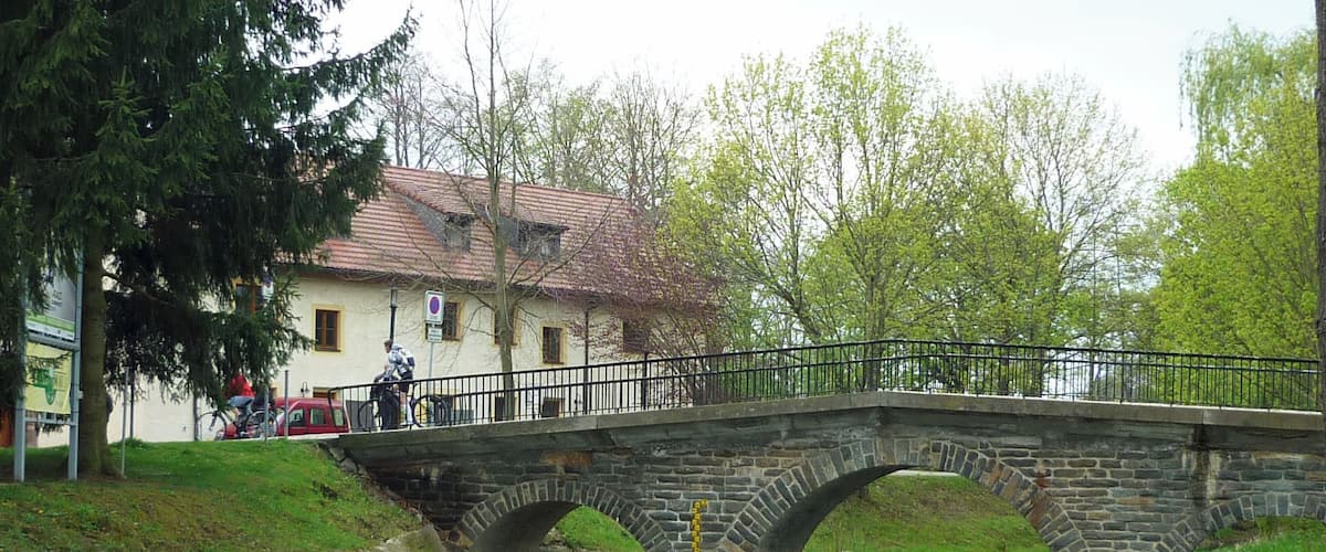 A stone bridge over a flowing river, surrounded by greenery and a building with a tiled roof.