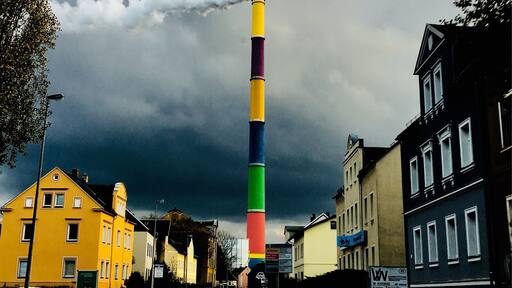 Un chimenea alta y colorida que emite humo, rodeada de edificios y un cielo nublado.