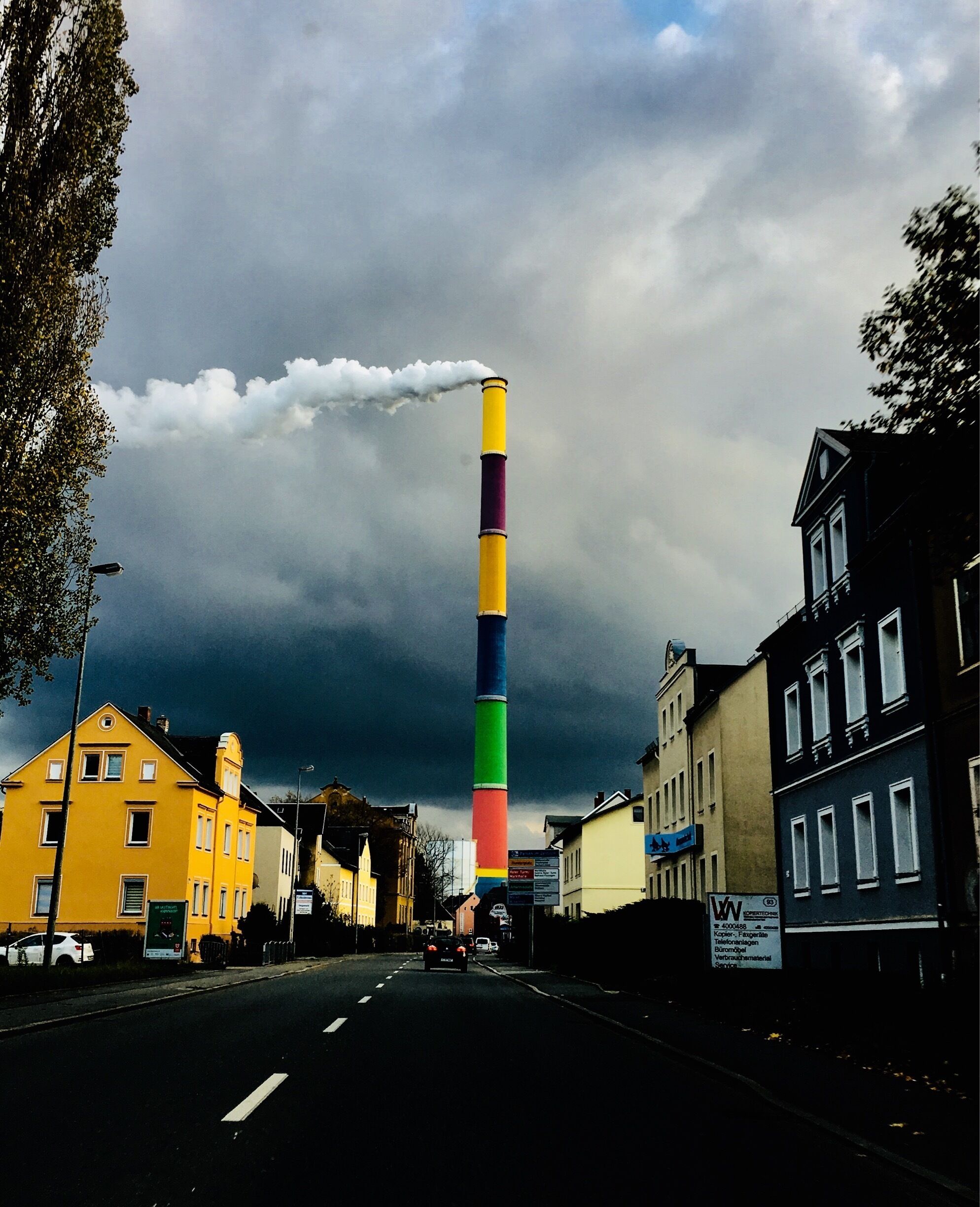 A tall, colorful chimney emitting smoke, surrounded by buildings and a cloudy sky.