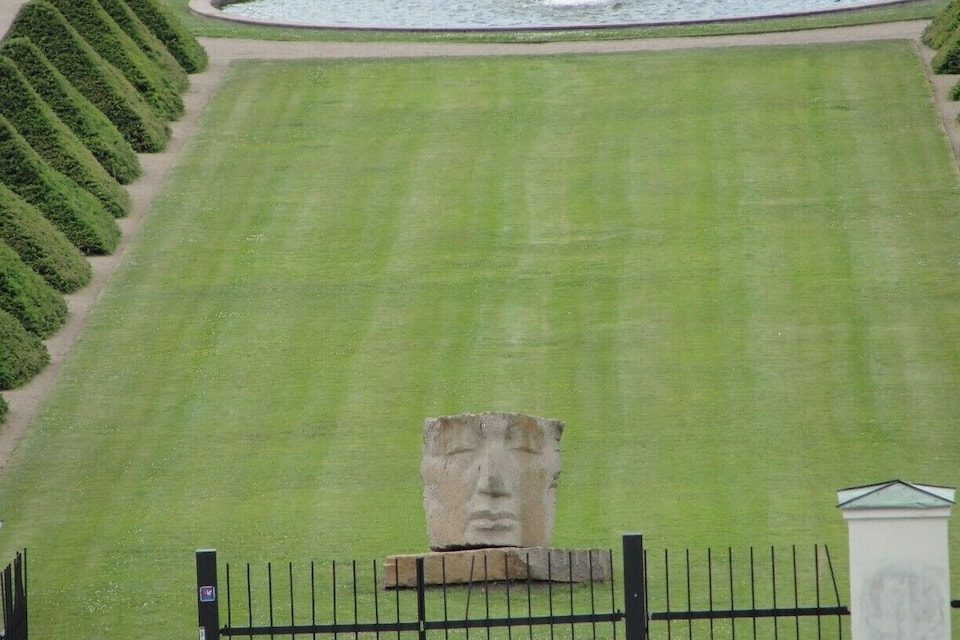 A well-manicured lawn with a stone statue in the foreground and a circular fountain in the background.