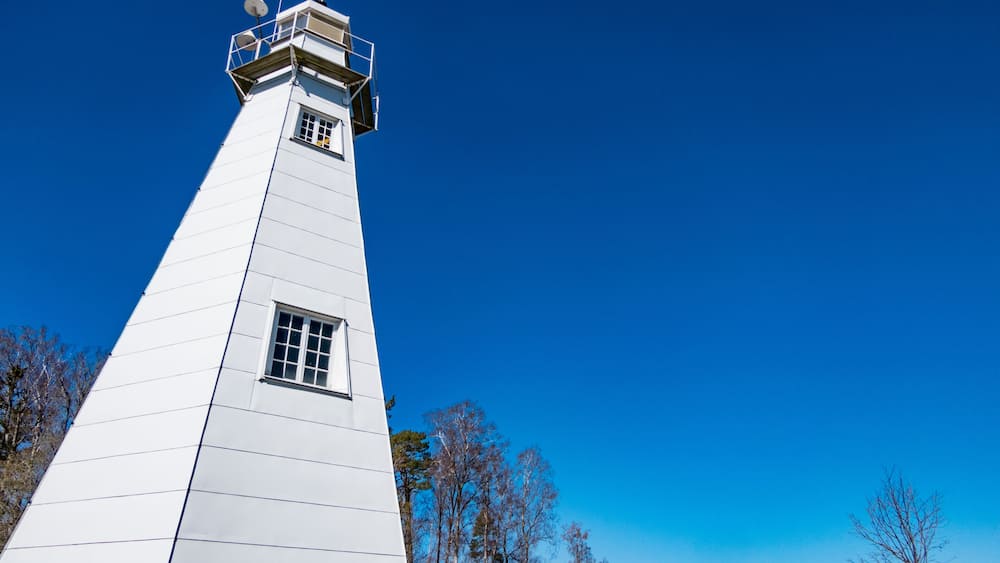 A tall white lighthouse with a small window and a balcony, surrounded by trees and a clear blue sky.