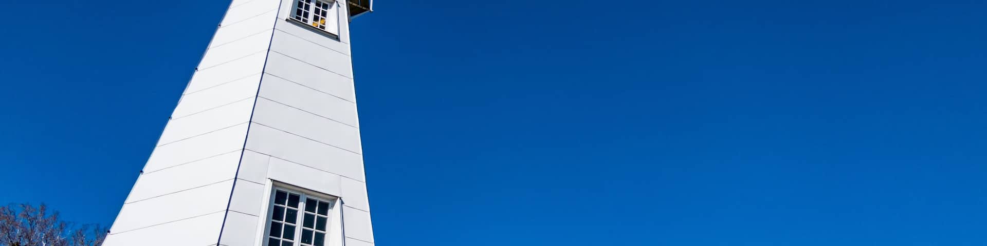 A tall white lighthouse with a small window and a balcony, surrounded by trees and a clear blue sky.