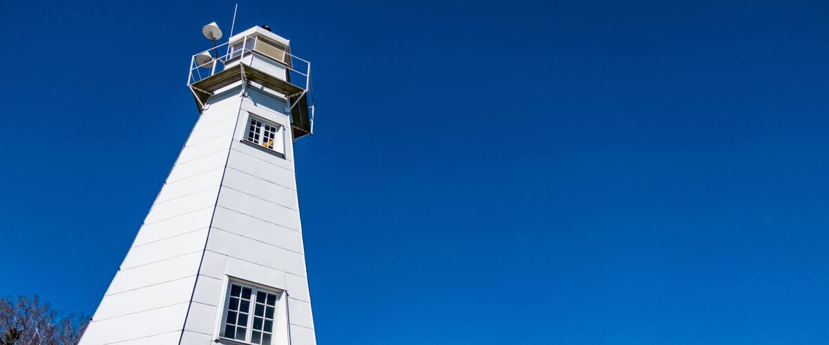 A tall white lighthouse with a small window and a balcony, surrounded by trees and a clear blue sky.