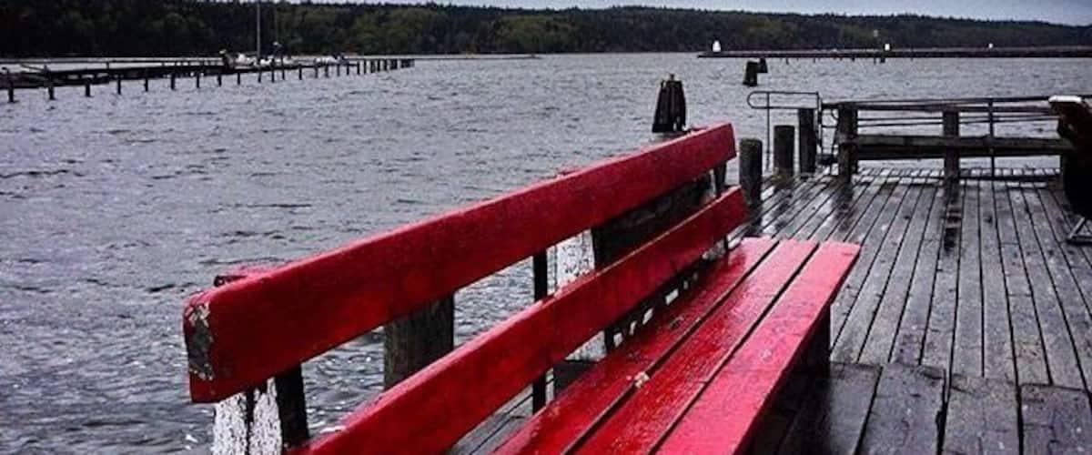 A red bench on a wooden pier extending into a body of water, with a cloudy sky above.