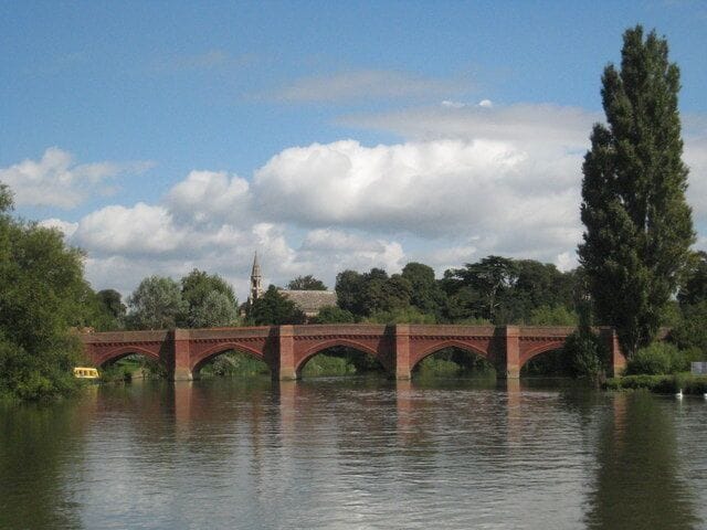 A red brick bridge over a calm river with trees and a distant church spire.